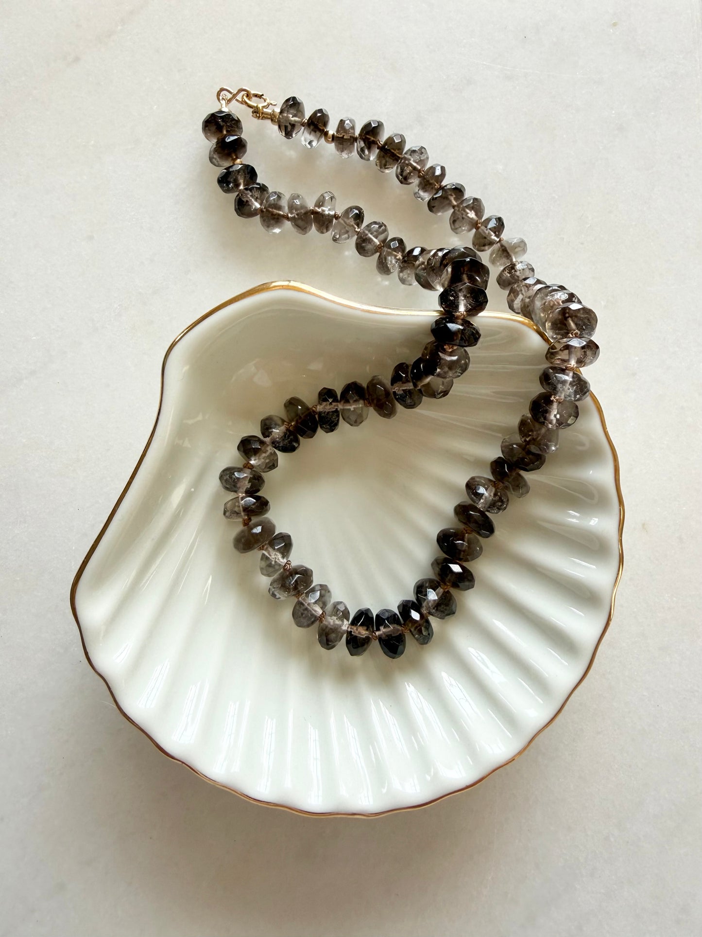 A hand knotted necklace made of faceted smoky quartz beads, draped over a white scalloped shell-shaped dish with a gold rim on a light marble surface.