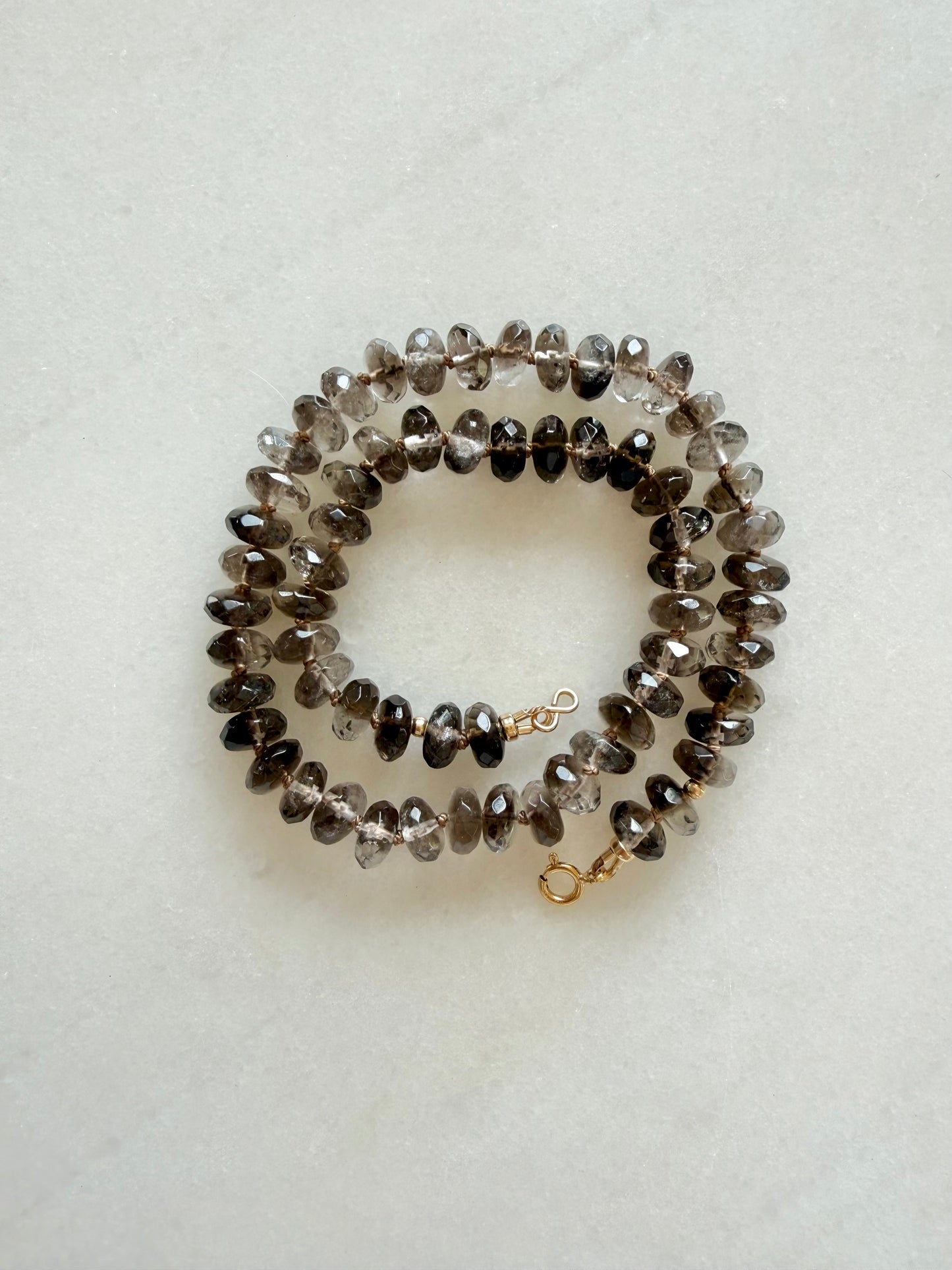 A hand-knotted necklace made of faceted smoky gray and brown gemstone beads with a small gold clasp, displayed on a light textured background.