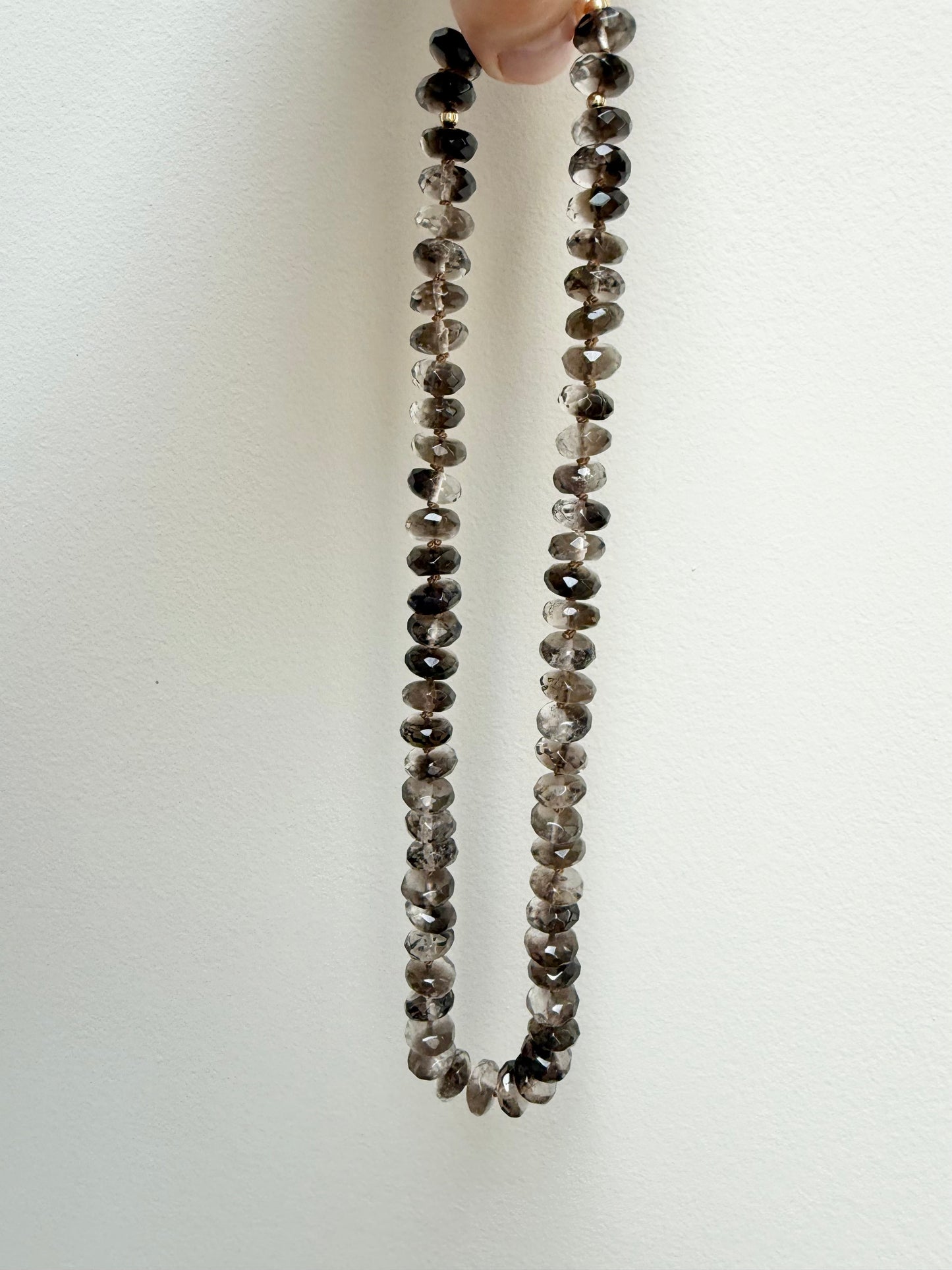 A beaded necklace made of small round gray and brown gemstone beads, held up by a hand against a plain off-white background.