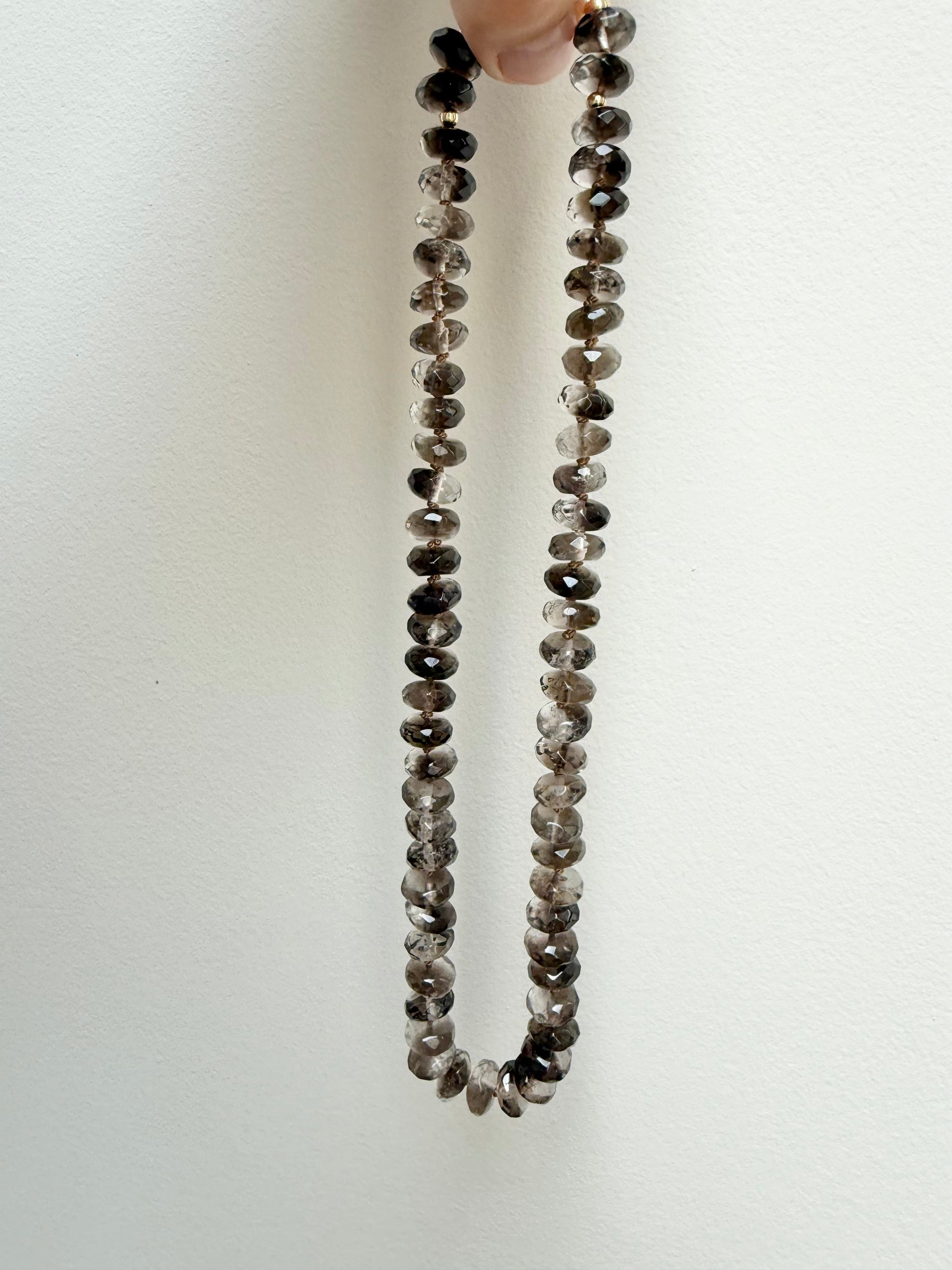 A beaded necklace made of small round gray and brown gemstone beads, held up by a hand against a plain off-white background.