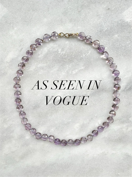 Purple beaded necklace on a textured gray background with 'As Seen in Vogue' text.