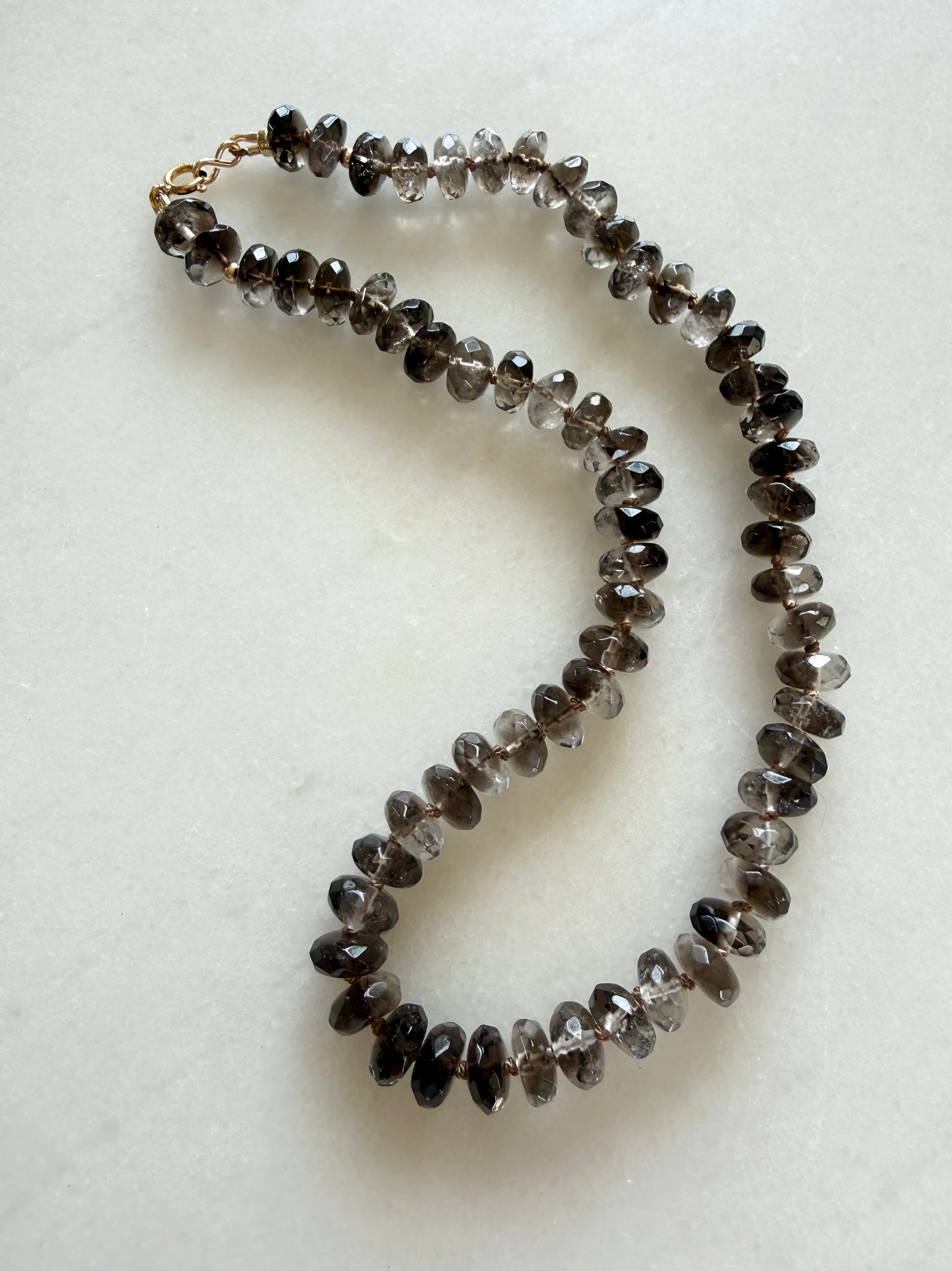 A hand knotted necklace made of faceted smoky quartz beads in shades of gray and brown, with a gold filled clasp, displayed on a light off-white textured surface.