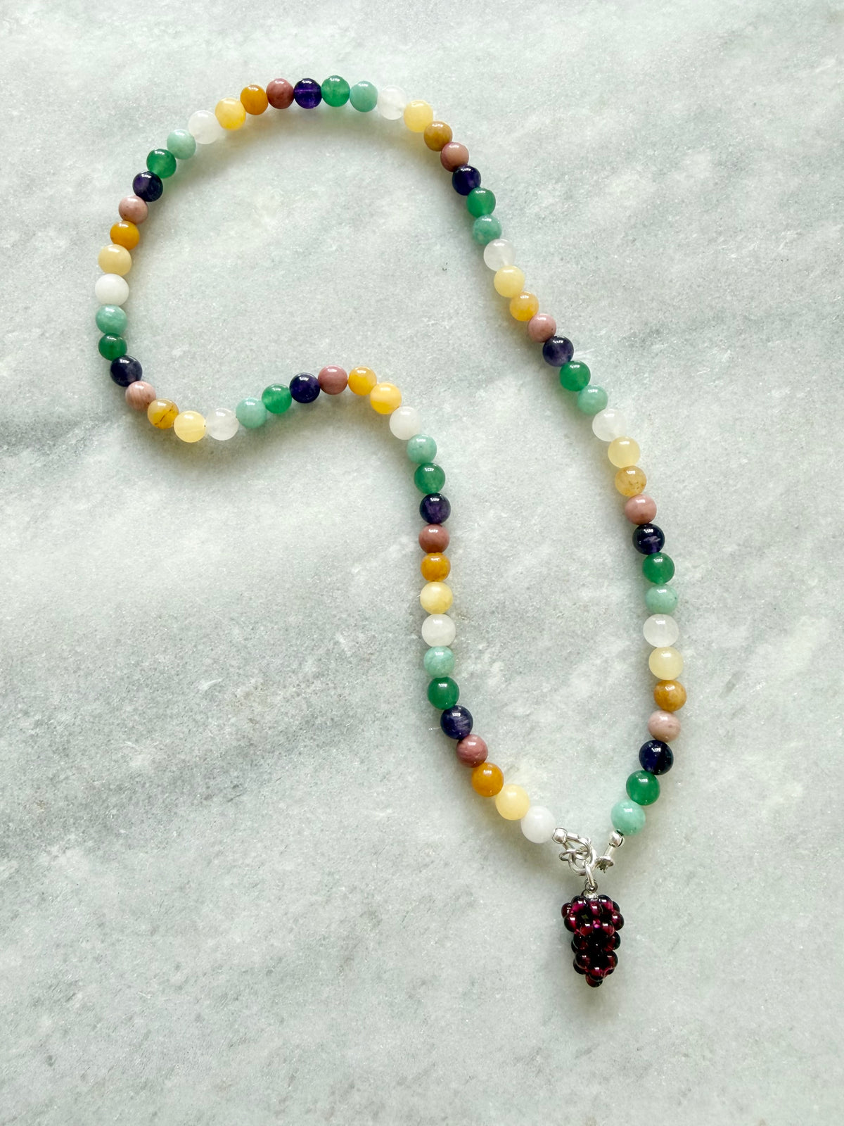 A beaded necklace with multicolored round beads in shades of green, yellow, white, purple, pink, and orange, featuring a dark red beaded grape cluster pendant on a silver connector, displayed on a light gray textured surface.