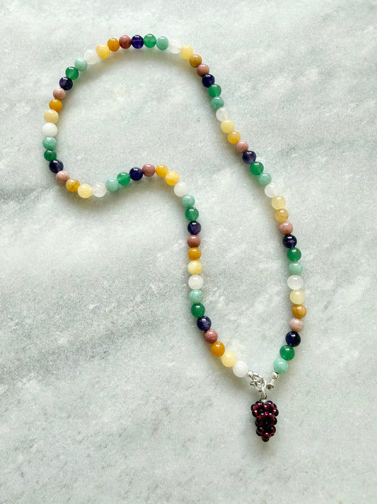 A beaded necklace with multicolored round beads in shades of green, yellow, white, purple, pink, and orange, featuring a dark red beaded grape cluster pendant on a silver connector, displayed on a light gray textured surface.