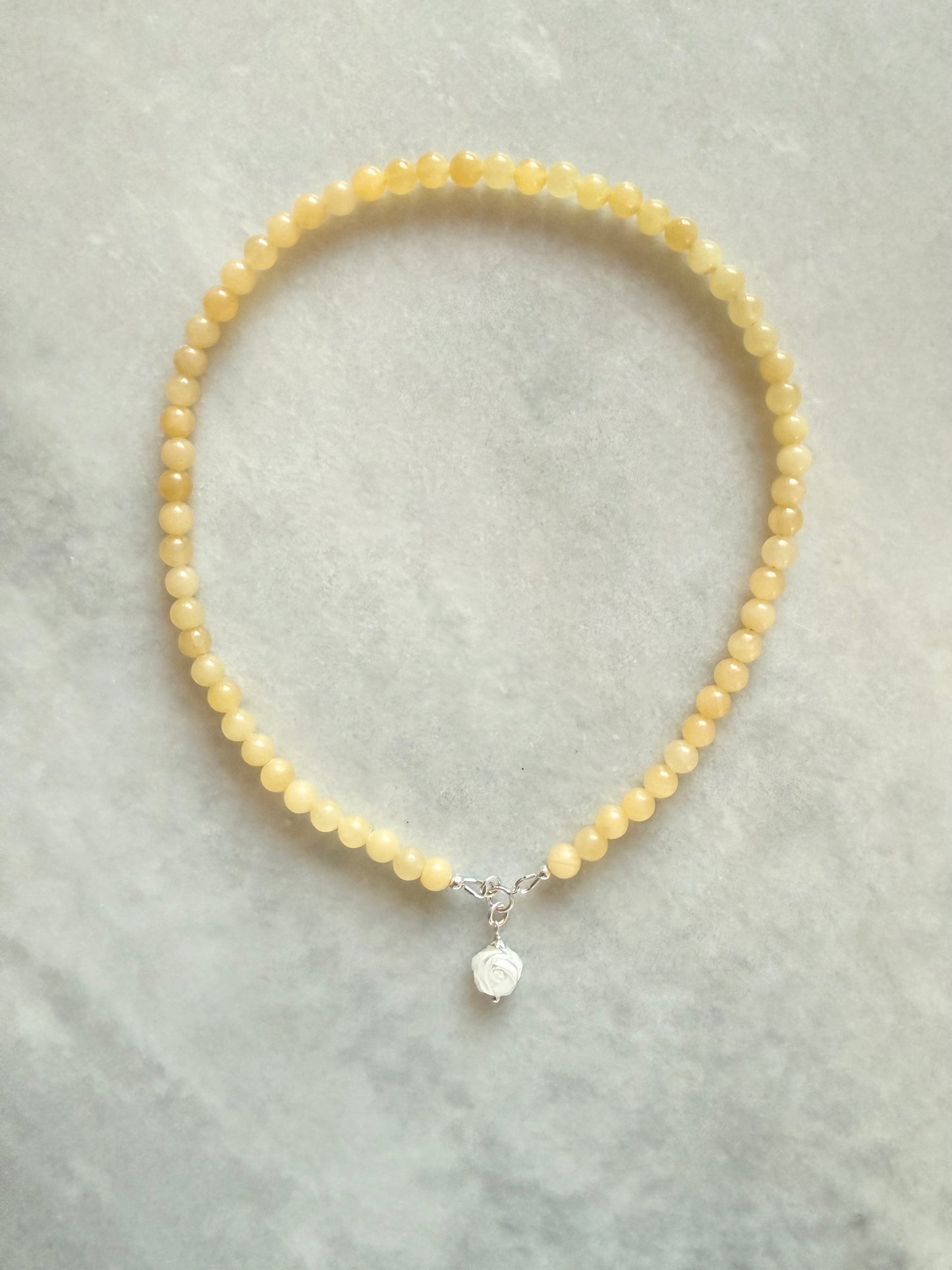 A delicate beaded necklace made of yellow butter quartz beads, featuring a sterling silver clasp with a mother of pearl rose charm, displayed on a light grey textured surface.