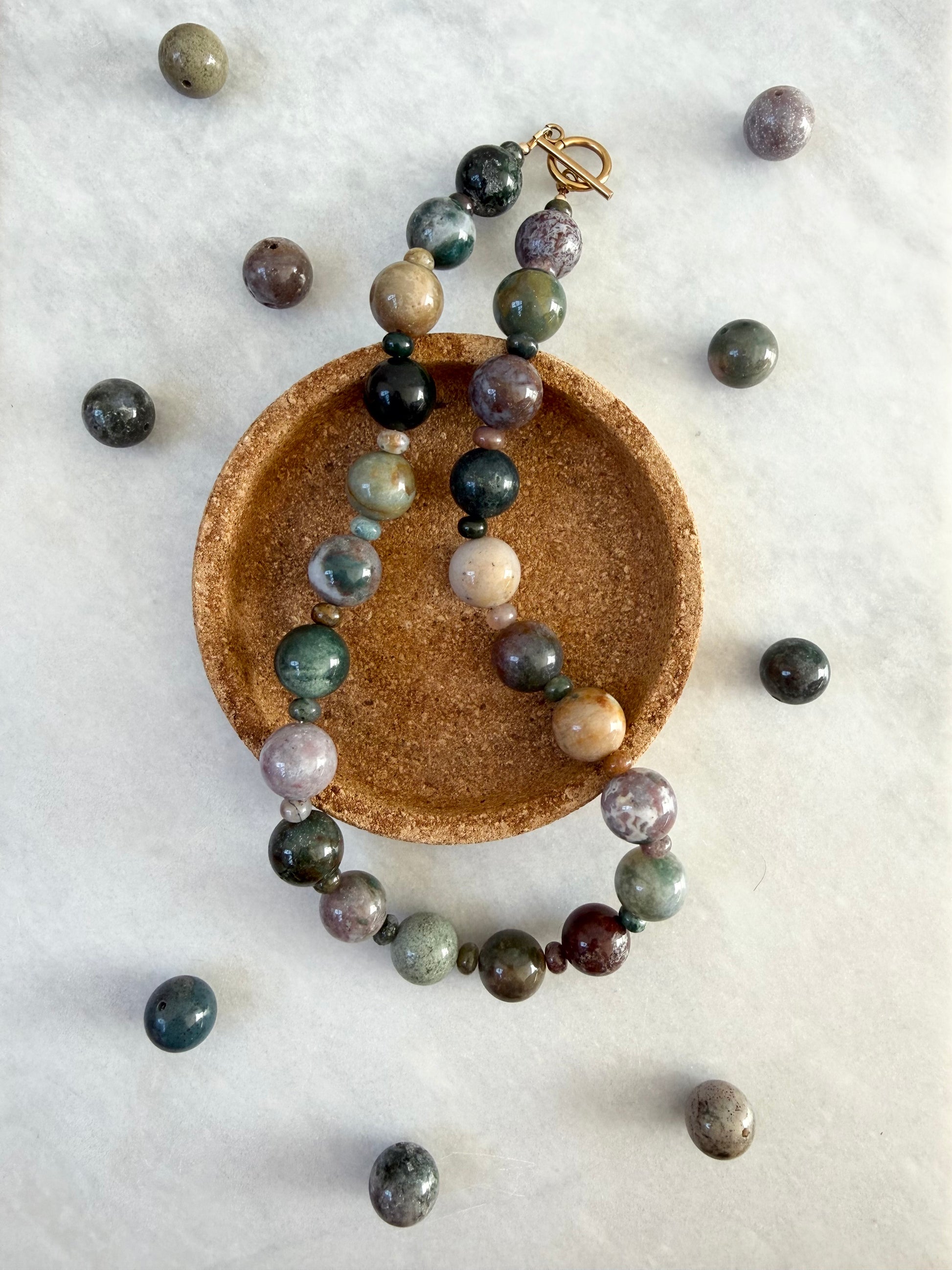 A beaded necklace made of polished multi-colored gemstone beads in shades of green, gray, purple, and beige, featuring a gold-plated toggle clasp. The necklace is draped over a round brown cork dish on a white textured background, with several loose beads scattered around it.