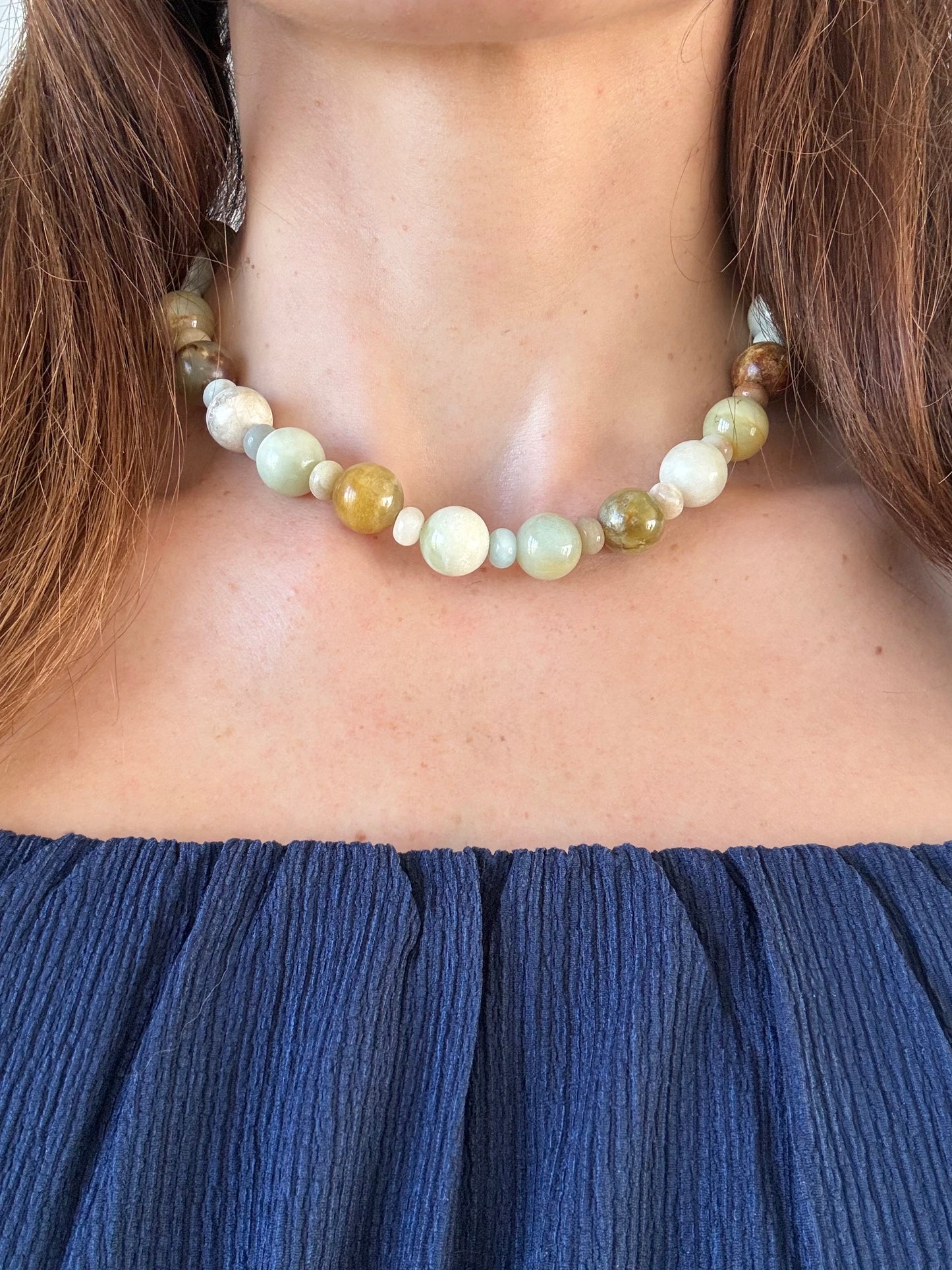 A close-up of a woman wearing a beaded necklace made of natural stone beads in muted green, cream, and brown tones, paired with a dark blue off-the-shoulder top.
