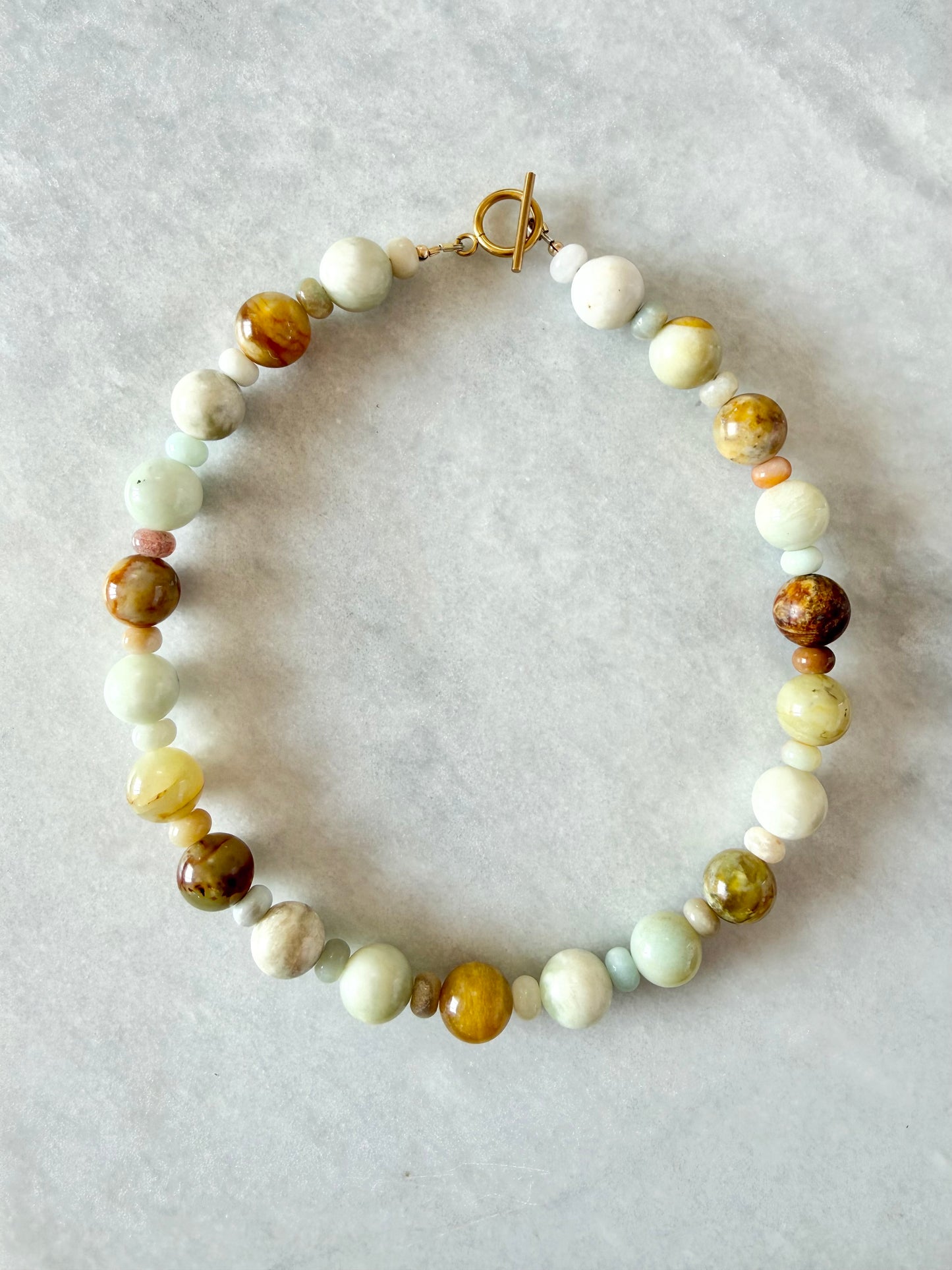 A handmade beaded necklace with a toggle clasp, featuring a mix of light green, cream, brown, and yellow stone beads, laid flat on a light gray textured background.