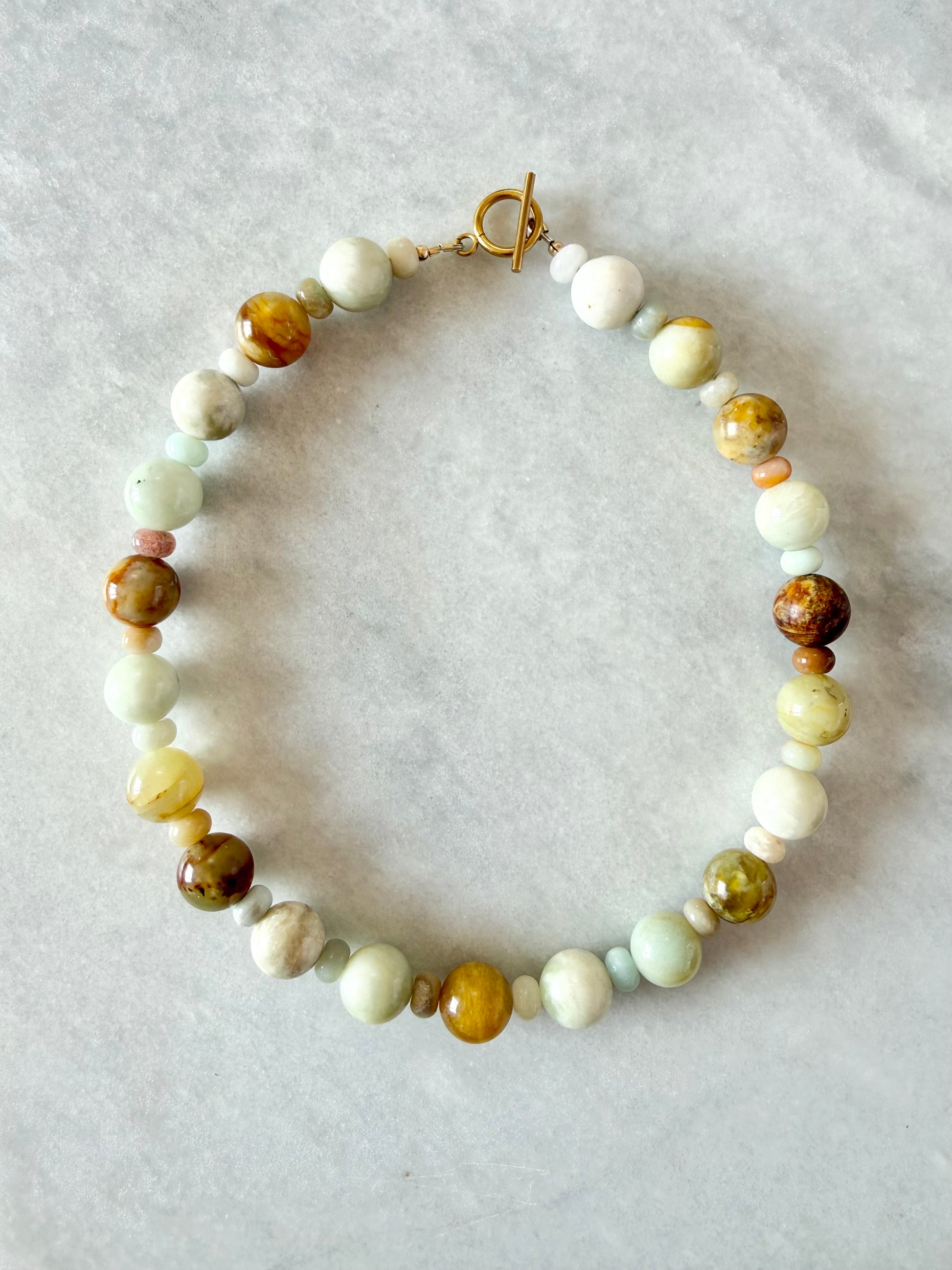A handmade beaded necklace with a toggle clasp, featuring a mix of light green, cream, brown, and yellow stone beads, laid flat on a light gray textured background.