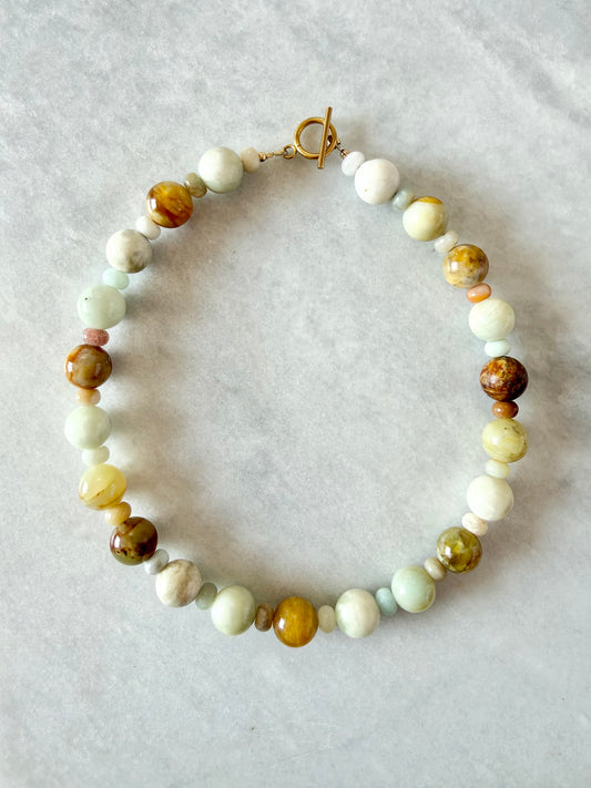 A handmade beaded necklace with a toggle clasp, featuring a mix of light green, cream, brown, and yellow stone beads, laid flat on a light gray textured background.