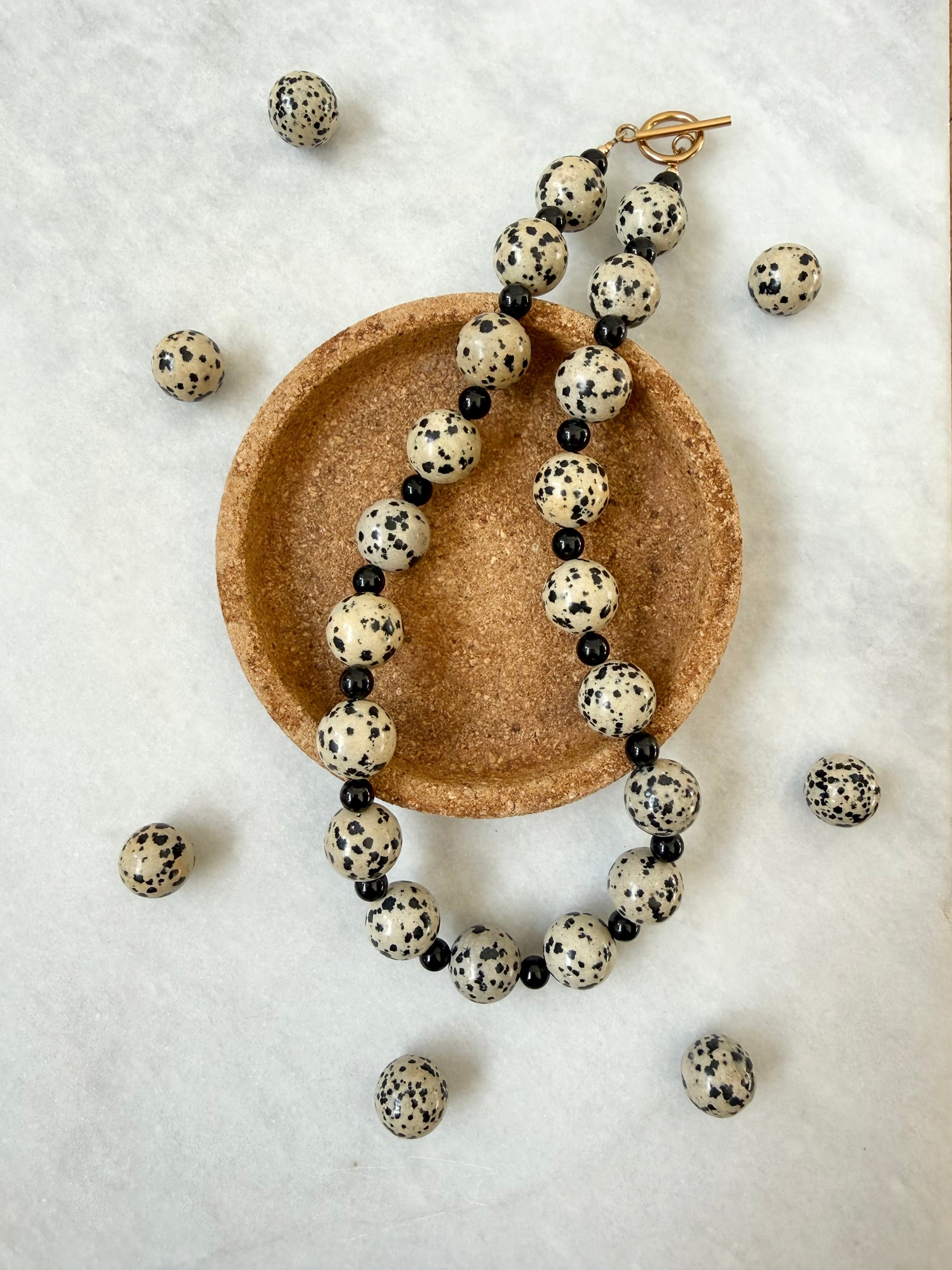 A beaded necklace made of round black-and-cream dalmatian jasper beads and small tourmaline beads, displayed on a round cork tray with a gold clasp, surrounded by scattered matching beads on a white textured background.