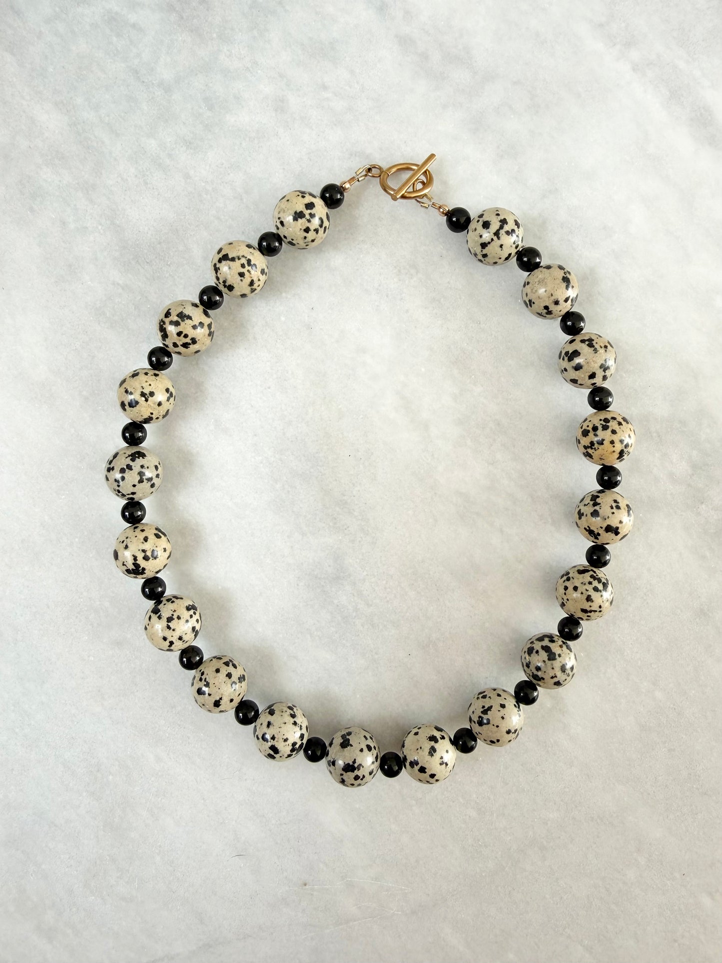 A beaded necklace featuring large round cream beads with black speckles, alternating with small black tourmaline beads, and finished with a gold-plated toggle clasp, displayed on a textured marble background.