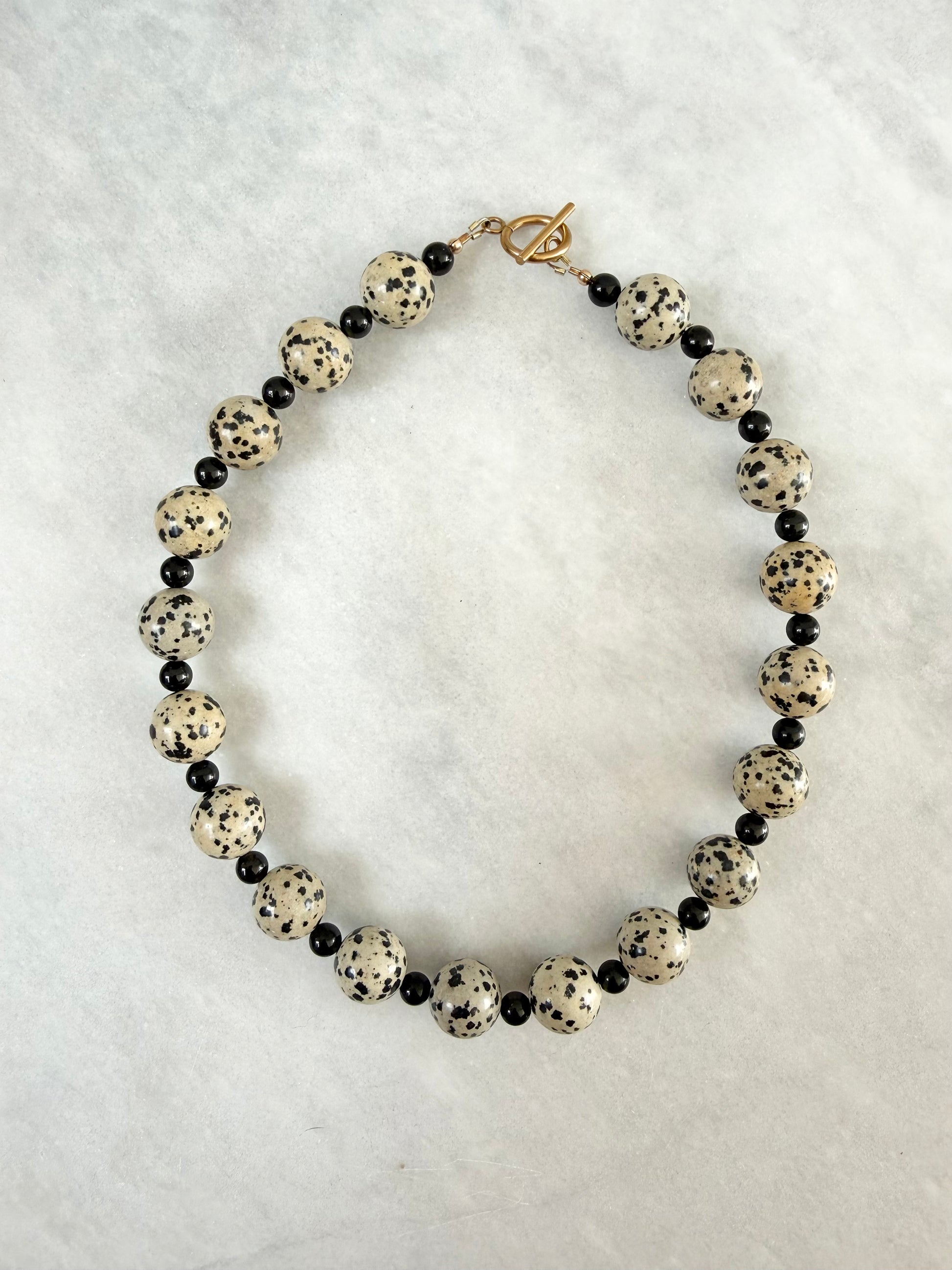 A beaded necklace featuring large round cream beads with black speckles, alternating with small black tourmaline beads, and finished with a gold-plated toggle clasp, displayed on a textured marble background.