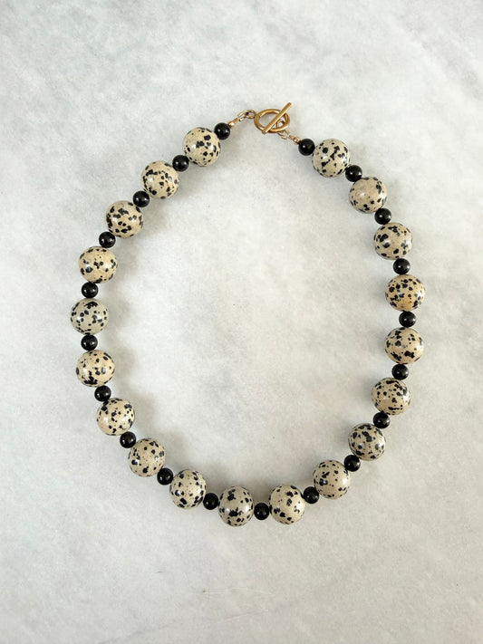 A beaded necklace featuring large round cream beads with black speckles, alternating with small black tourmaline beads, and finished with a gold-plated toggle clasp, displayed on a textured marble background.