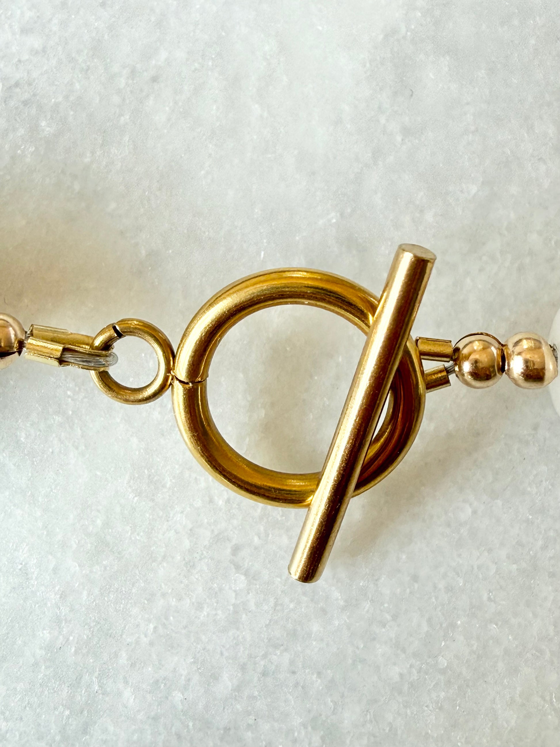 A close-up of a gold plated toggle clasp resting on a textured white surface.