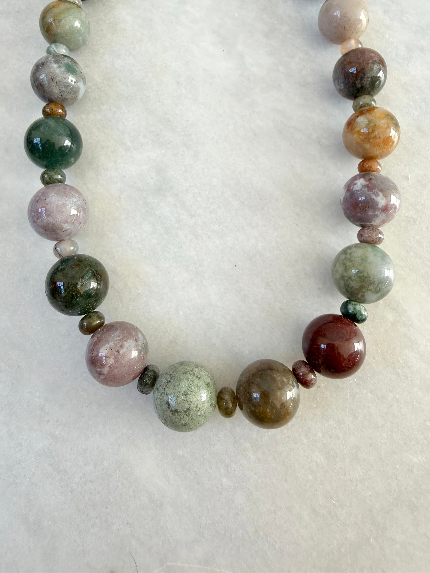 A beaded necklace made of polished gemstone beads in various earthy colors including green, pink, brown, and red, arranged in a U-shape on a marble background.
