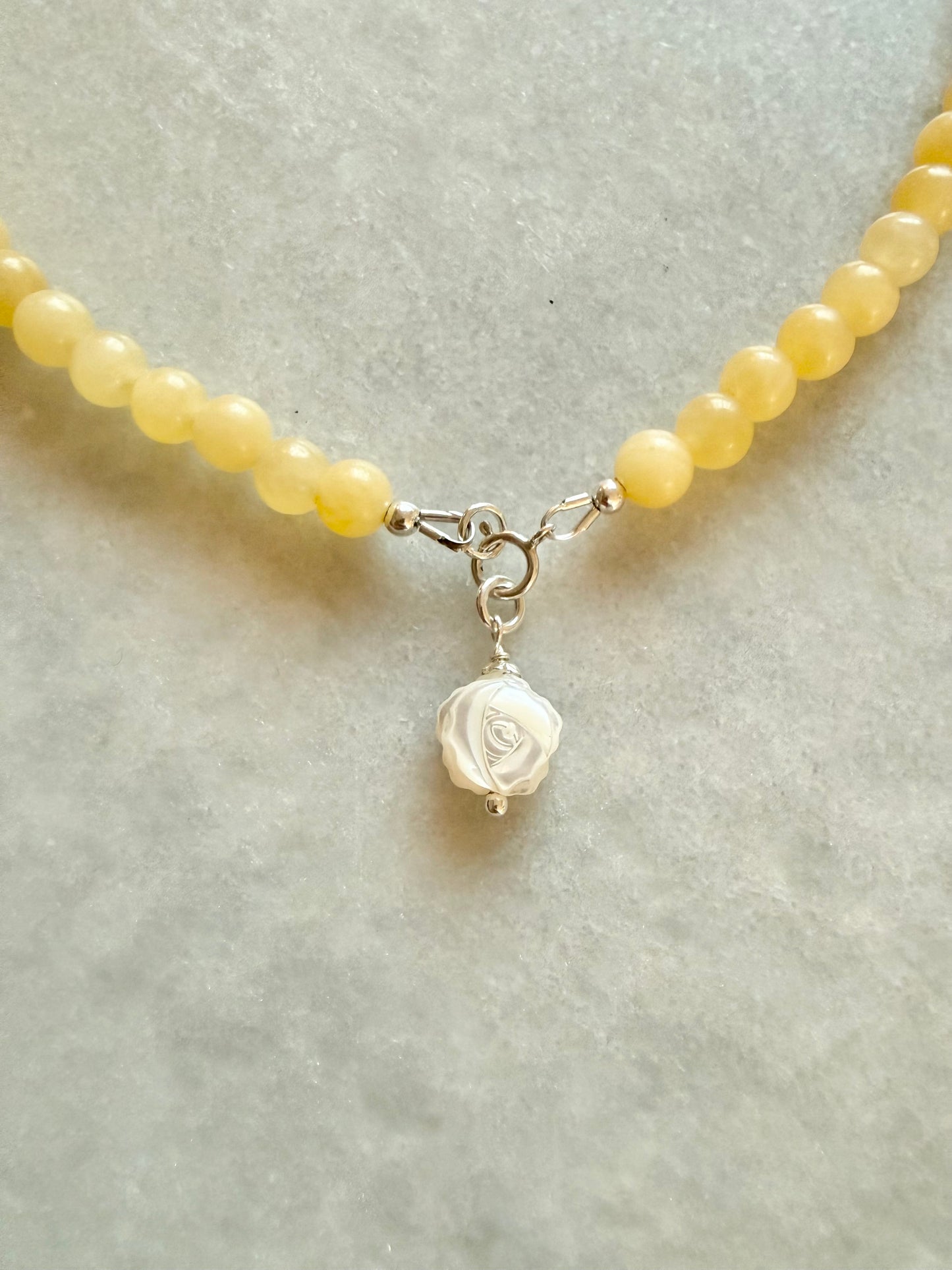 A close-up of a beaded necklace with yellow quartz round beads and a sterling silver clasp holding a white rose-shaped pendant, displayed on a light gray textured surface.