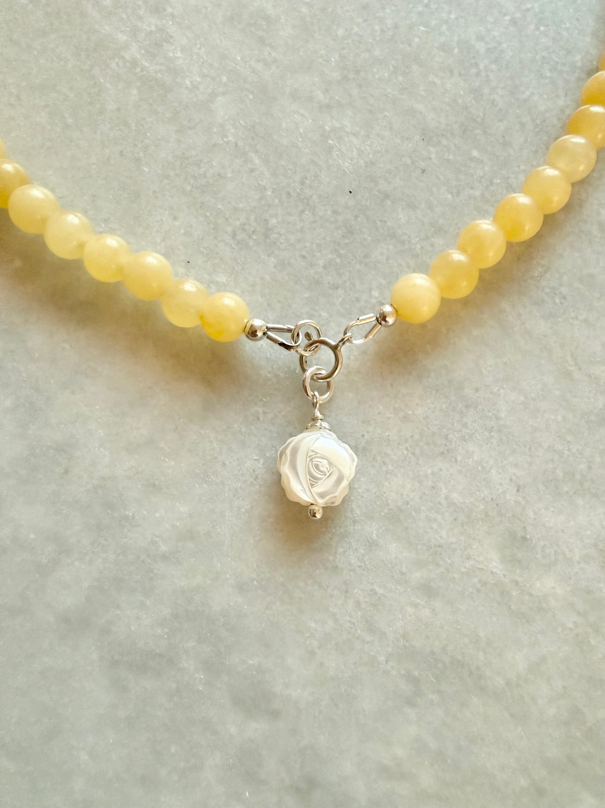 A close-up of a beaded necklace with yellow quartz round beads and a sterling silver clasp holding a white rose-shaped pendant, displayed on a light gray textured surface.
