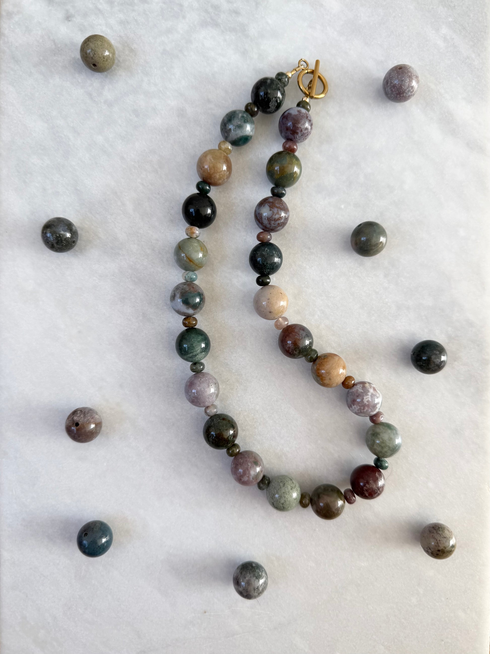 A beaded necklace made of polished gemstone beads in various earthy colors, arranged on a white marble surface with loose matching beads scattered around.