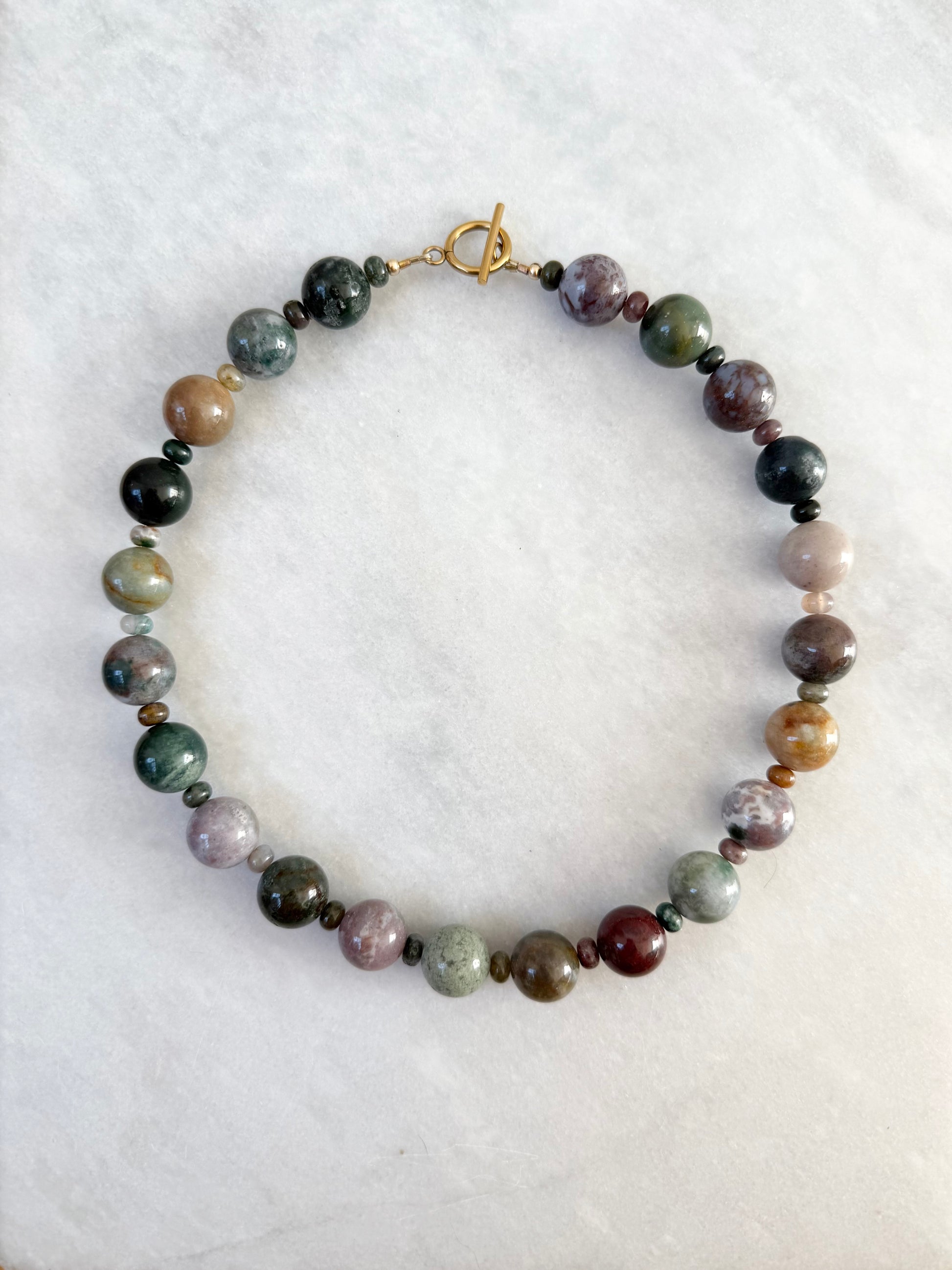 A beaded necklace made of polished round gemstone beads in various earthy colors, including green, brown, purple, and gray, separated by small dark spacer beads and finished with a gold-tone toggle clasp, displayed on a white marble background.