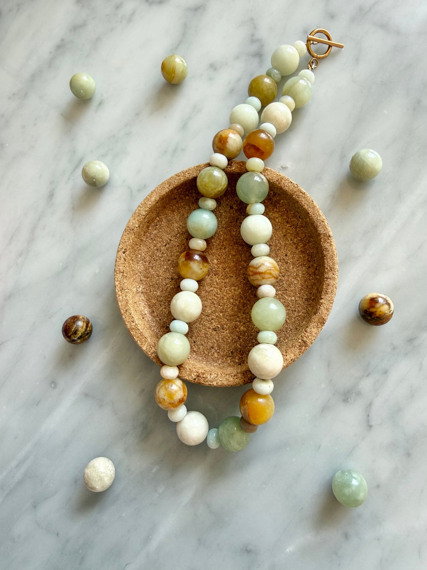 A handmade beaded necklace with a toggle clasp, displayed on a small round cork dish over a light gray marble surface. The beads are made of various stone types including light green jade and amazonite.