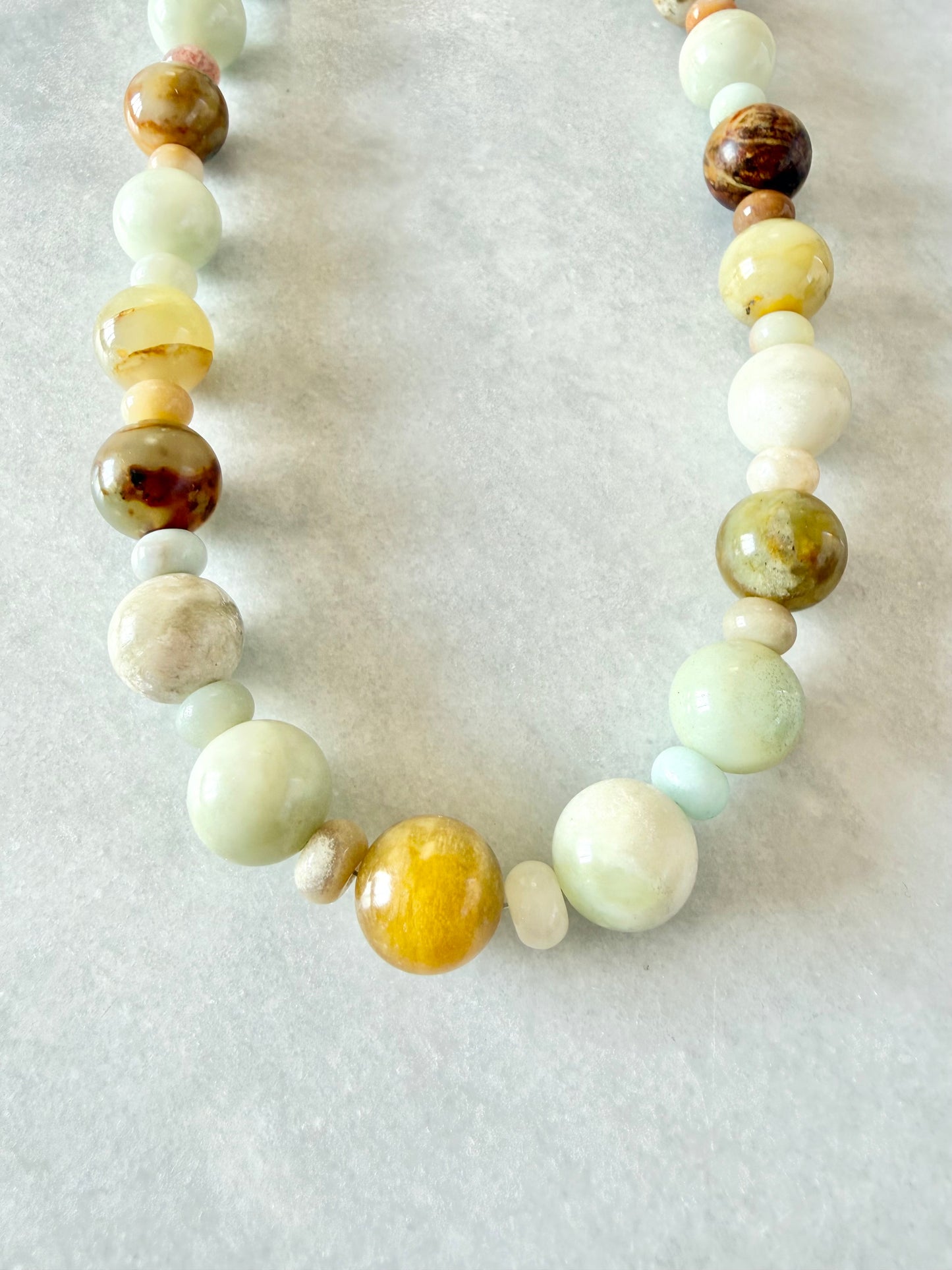 A beaded necklace made of polished round gemstone beads in soft pastel shades of mint green, pale yellow, cream, and brown, displayed on a light gray textured surface.