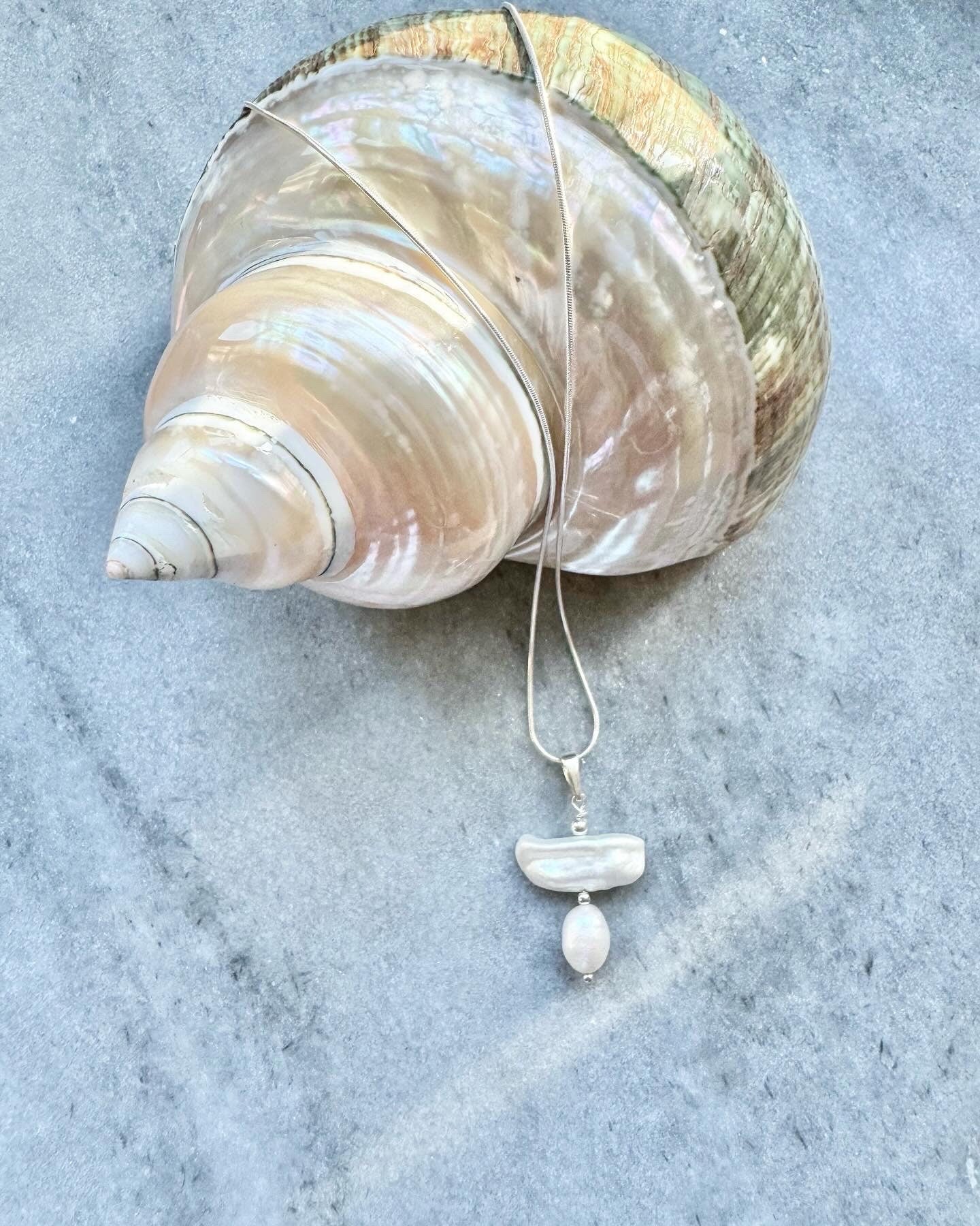 A sterling silver snake chain necklace with a biwa pearl silver pendant, displayed on a marble surface next to a large seashell.